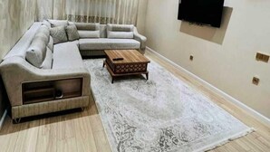 Ein Wohnzimmer mit einer Eckcouch, einem Couchtisch, einem gemusterten Teppich und einem Flachbildfernseher an der Wand.