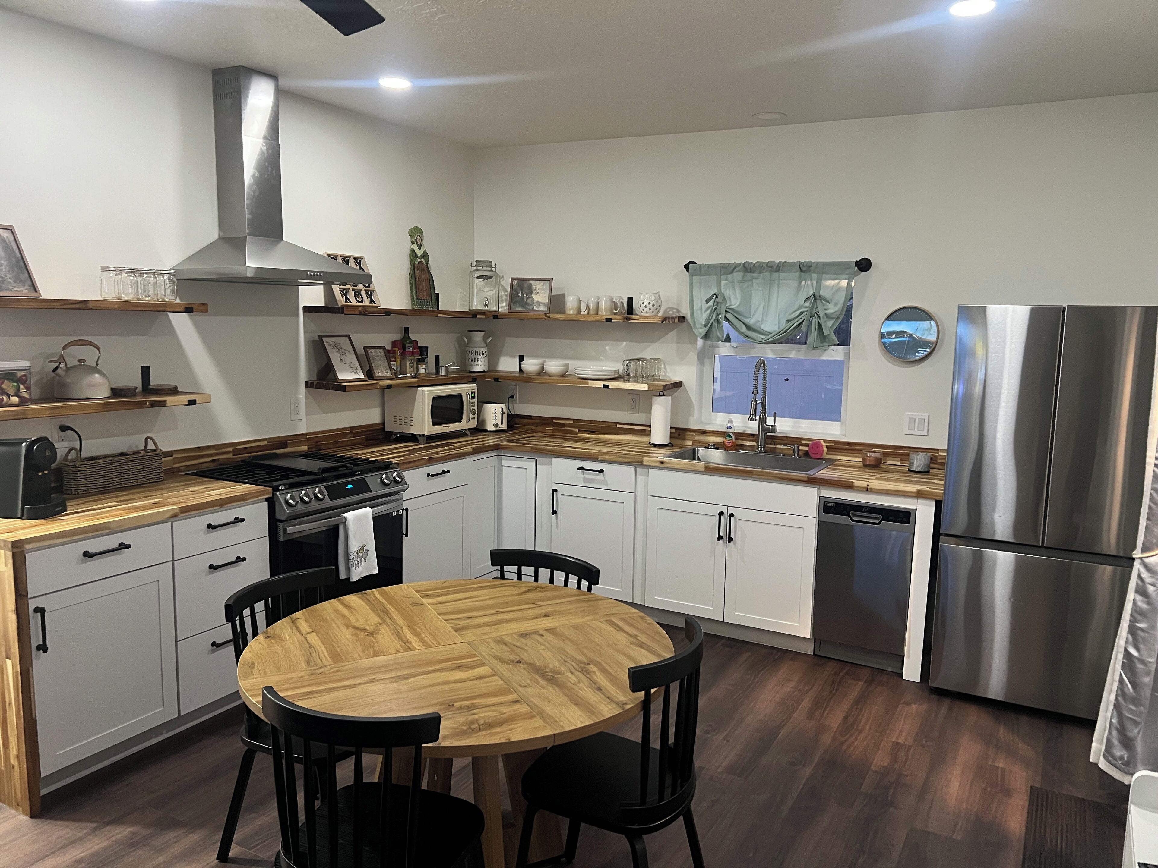 A modern kitchen with white cabinets, a stainless steel refrigerator, a microwave, and a stove. There is a wooden dining table with chairs.