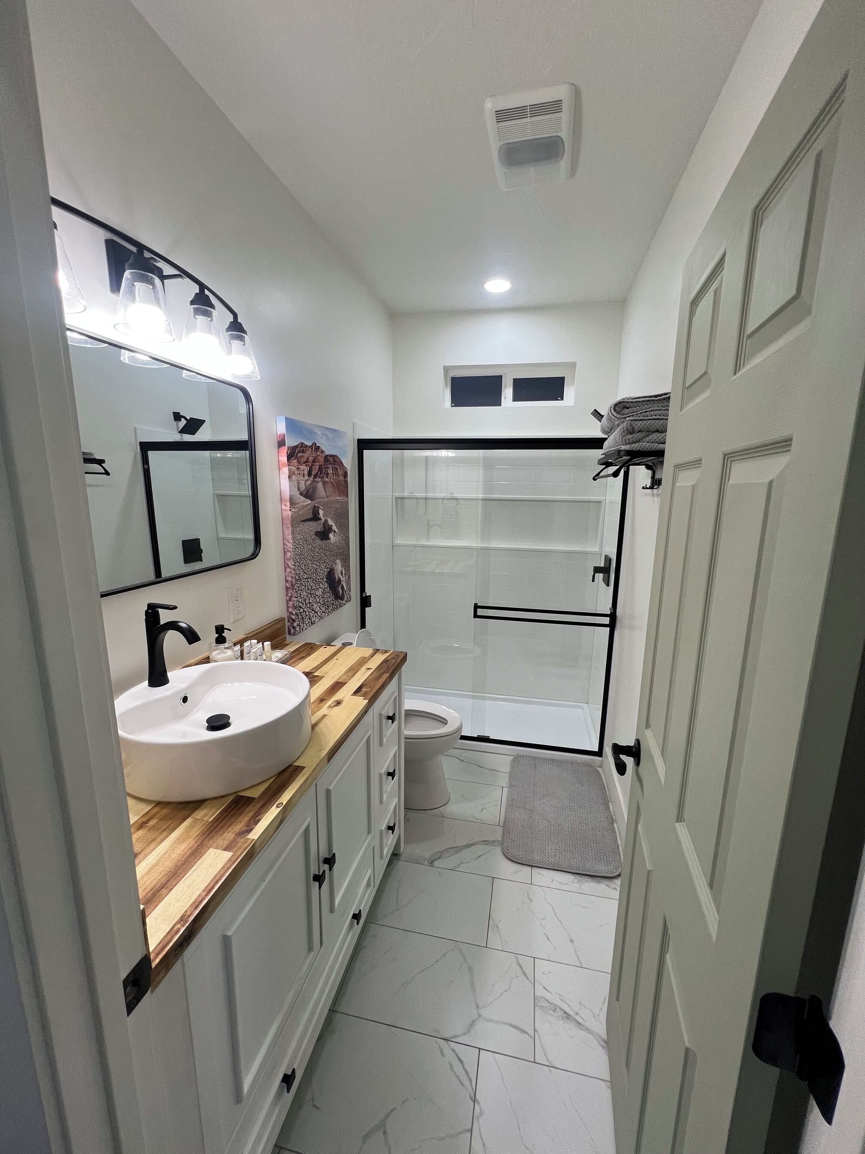 A bathroom with a white sink, wooden vanity, a mirror with lights, a toilet, and a shower area.