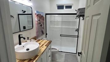 A bathroom with a white sink, wooden vanity, a mirror with lights, a toilet, and a shower area.