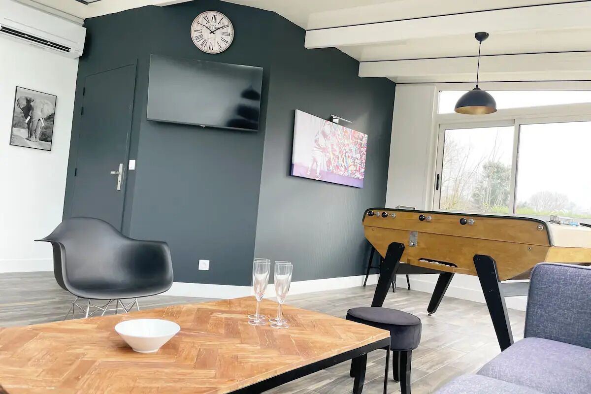 Un salon moderne avec une table en bois, deux chaises, un canapé, un téléviseur fixé au mur, une horloge et une grande fenêtre.