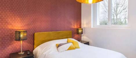 Habitación de hotel con pared de ladrillo, una cama grande con ropa de cama blanca y almohadas amarillas, dos lámparas de noche y una ventana con vistas a árboles.