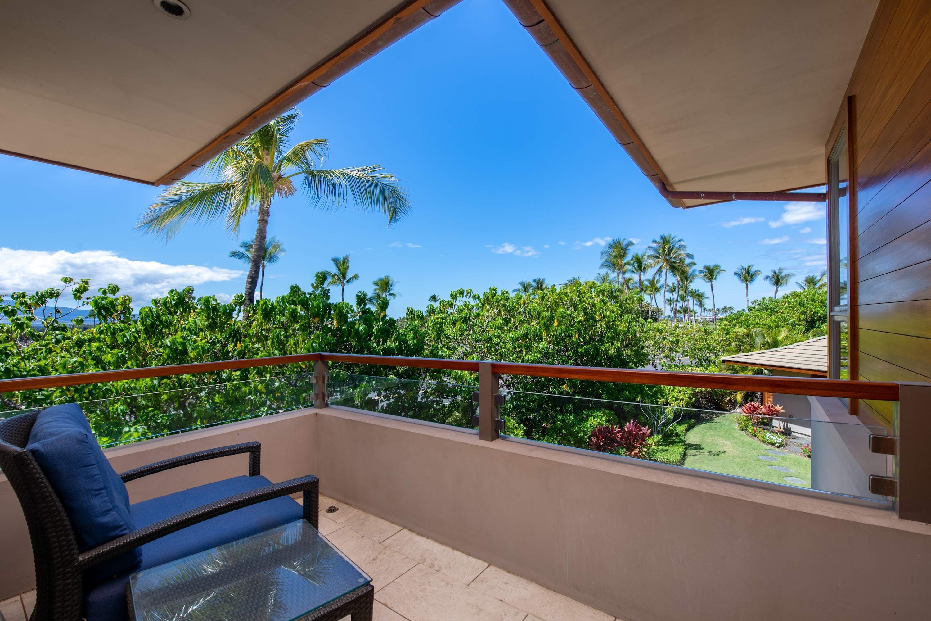 A balcony with a glass table and a chair, overlooking a lush garden and palm trees.