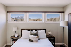 A bedroom with a large bed, three windows with mountain views, a bedside table with a lamp, and a nightstand with a vase of flowers.