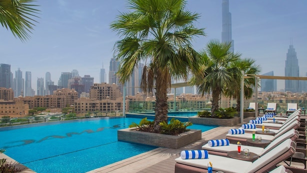 A rooftop pool with lounge chairs and palm trees, overlooking a cityscape with tall buildings. - Cozy 1BR with balcony Damac Towers (Dubai)