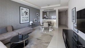 A modern living room with a sofa, coffee table, and TV. There is a dining area with chairs and a table. The kitchen is visible in the background. - Cozy 1BR with balcony Damac Towers (Dubai)