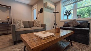 Ein Wohnzimmer mit Couchtisch aus Holz, zwei Sofas, einer Lampe und einem Fenster mit Blick auf Grünflächen.