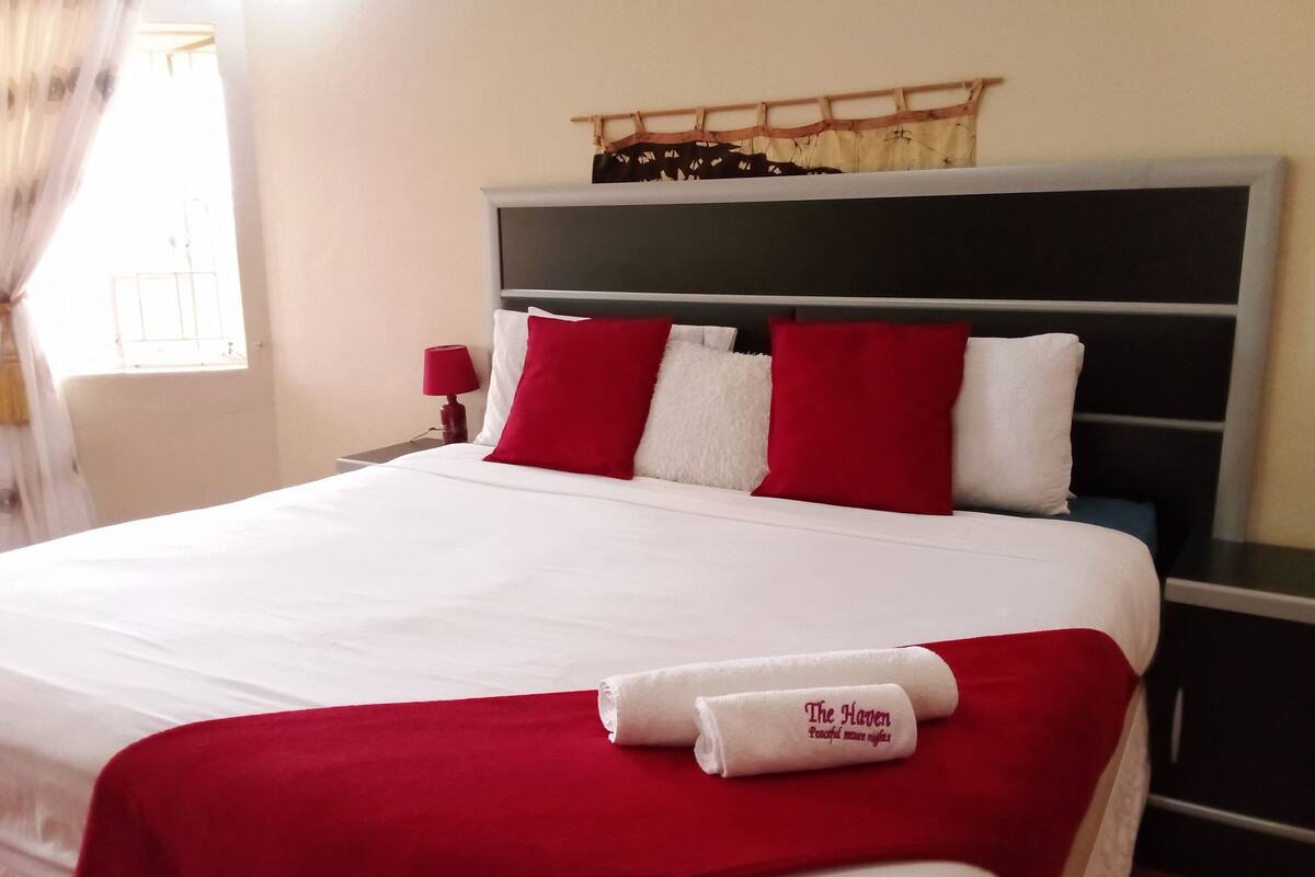 Een net opgemaakt bed met witte en rode beddengoed, een rode plaid en een opgerolde handdoek met tekst.