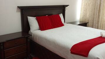 Una cama bien hecha con ropa de cama blanca y detalles rojos, una mesita de noche de madera oscura y una alfombra roja.
