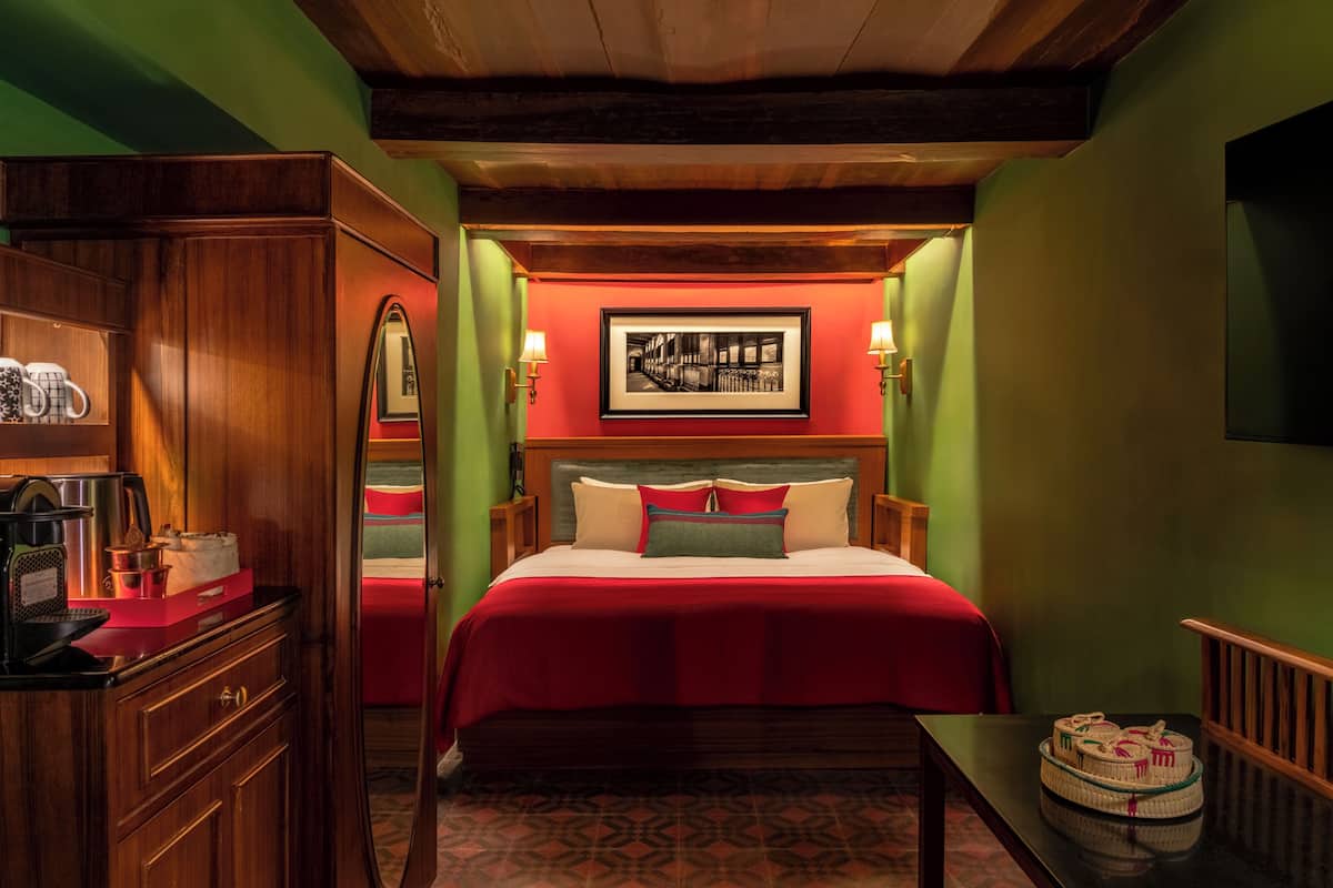 A hotel room with a wooden dresser, a bed with red bedding, a framed picture on the wall, and a small table with baskets.