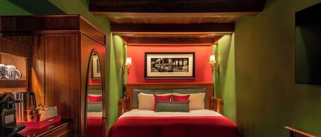 Ein Hotelzimmer mit einem Holzkasten, einem Bett mit roter Bettwäsche, einem gerahmten Bild an der Wand und einem kleinen Tisch mit Körben.