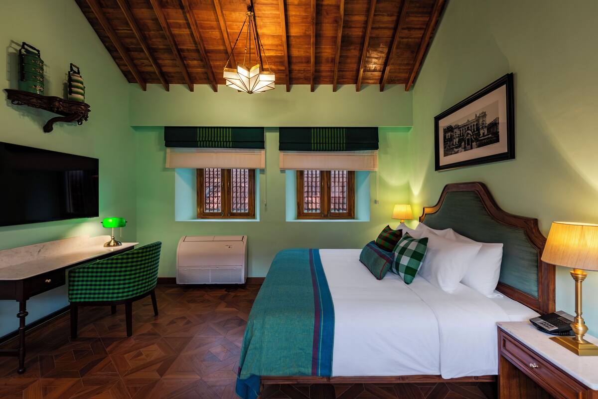 A hotel room with a wooden ceiling, a bed with a green and blue bedspread, a desk with a green chair, a television, and a framed picture on the wall.