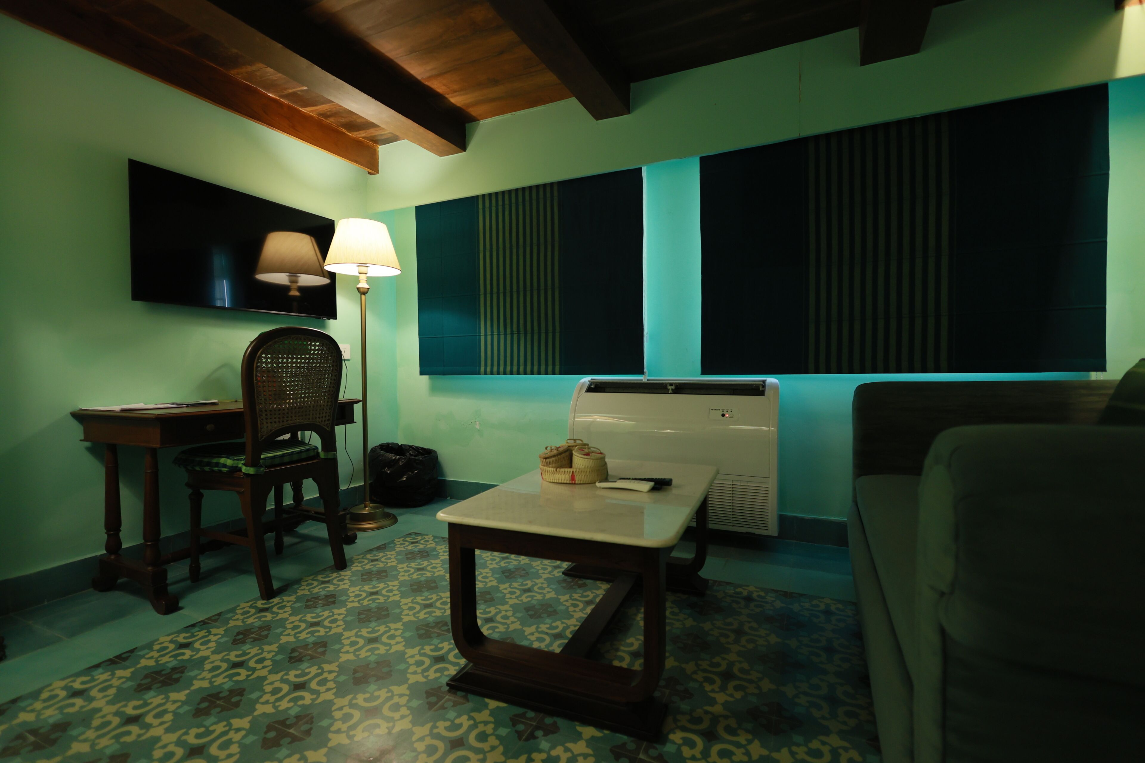 Une pièce avec un sol à motifs, un bureau avec une chaise, une table, une lampe, un téléviseur et une grande fenêtre avec des stores.