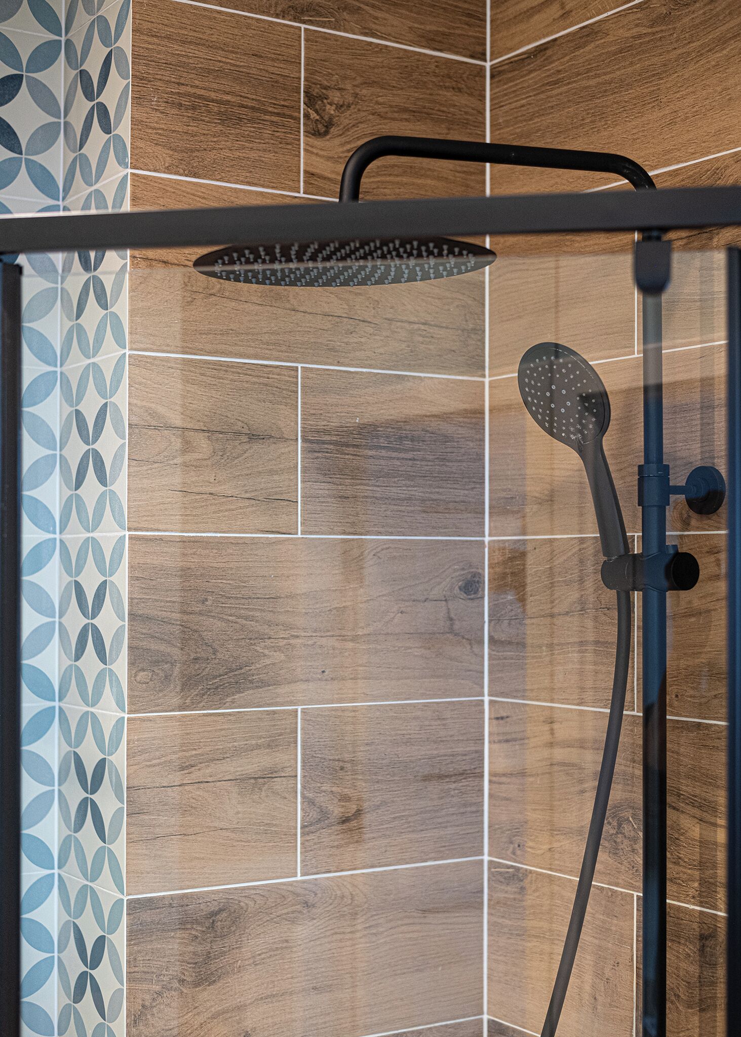 A shower enclosure with a rain showerhead and a handheld showerhead, set against a backdrop of wooden tiles and patterned tiles.