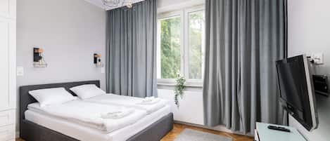 Een moderne slaapkamer met een groot bed, grijze gordijnen, een televisie en een raam met een plant.