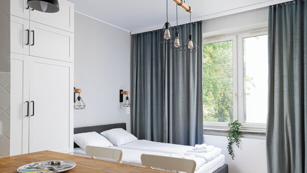 Een moderne slaapkamer met een bed, nachtkastjes, een raam met gordijnen en een zithoek met een tafel en stoelen.