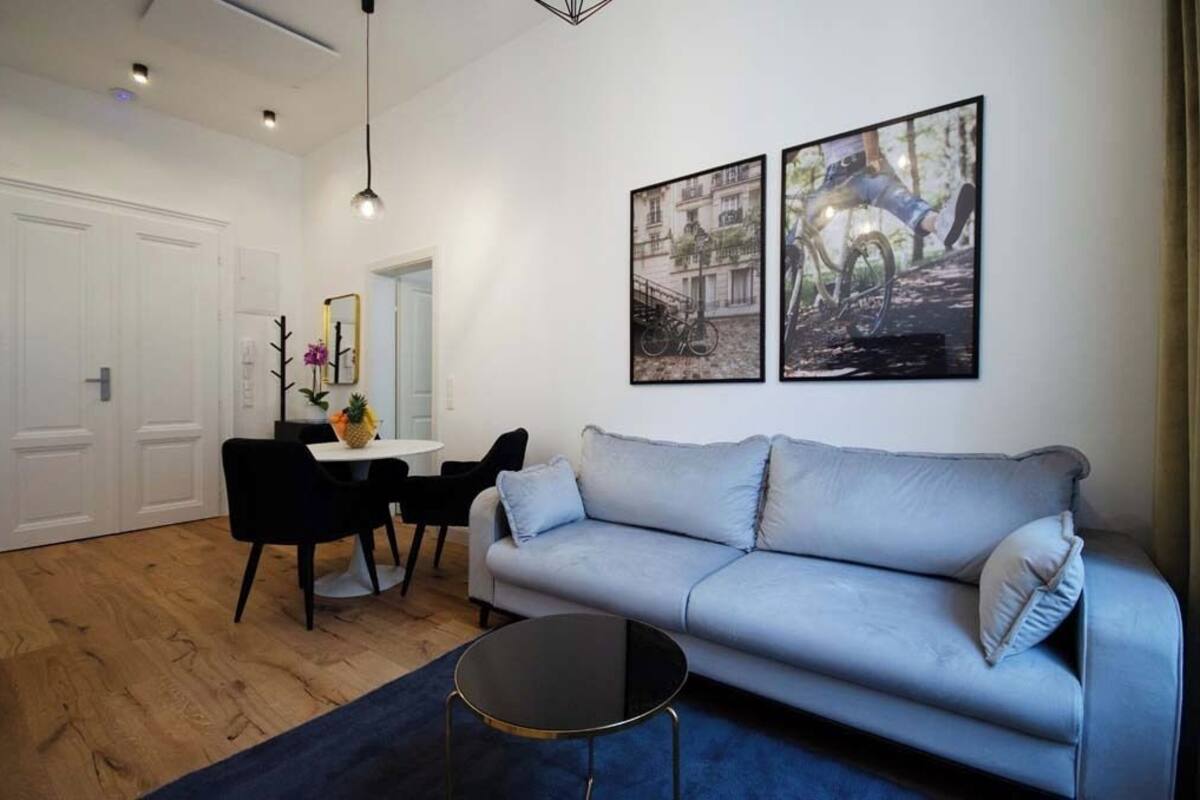 Een moderne woonkamer met een grijze bank, een ronde salontafel, en een eethoek met een witte tafel en zwarte stoelen. Twee ingekaderde foto’s hangen aan de muur en er is een deur in de achtergrond.