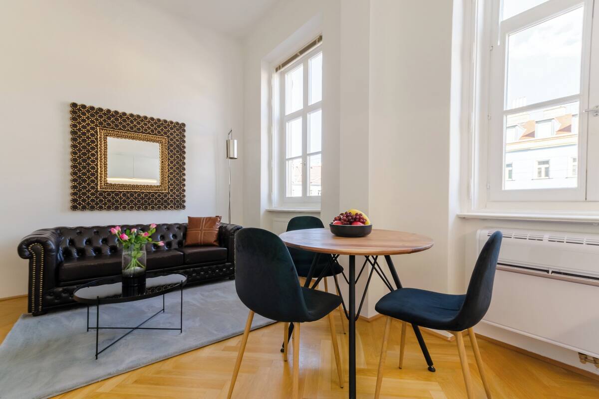 Een moderne woonkamer met een zwarte leren bank, een ronde houten tafel met stoelen, een decoratieve spiegel en een groot raam met uitzicht op gebouwen.