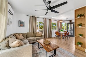 A living room with a sofa, a wooden coffee table, a dining area with chairs, a ceiling fan, and a bookshelf with plants. - Royal Point Modern Hale (Kailua Kona)