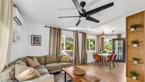 A living room with a sofa, a wooden coffee table, a dining area with chairs, a ceiling fan, and a bookshelf with plants.