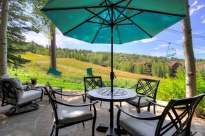 Relax in a serene outdoor setting with a round table and four comfortable chairs, shaded by a large umbrella amidst lush greenery and a scenic view.