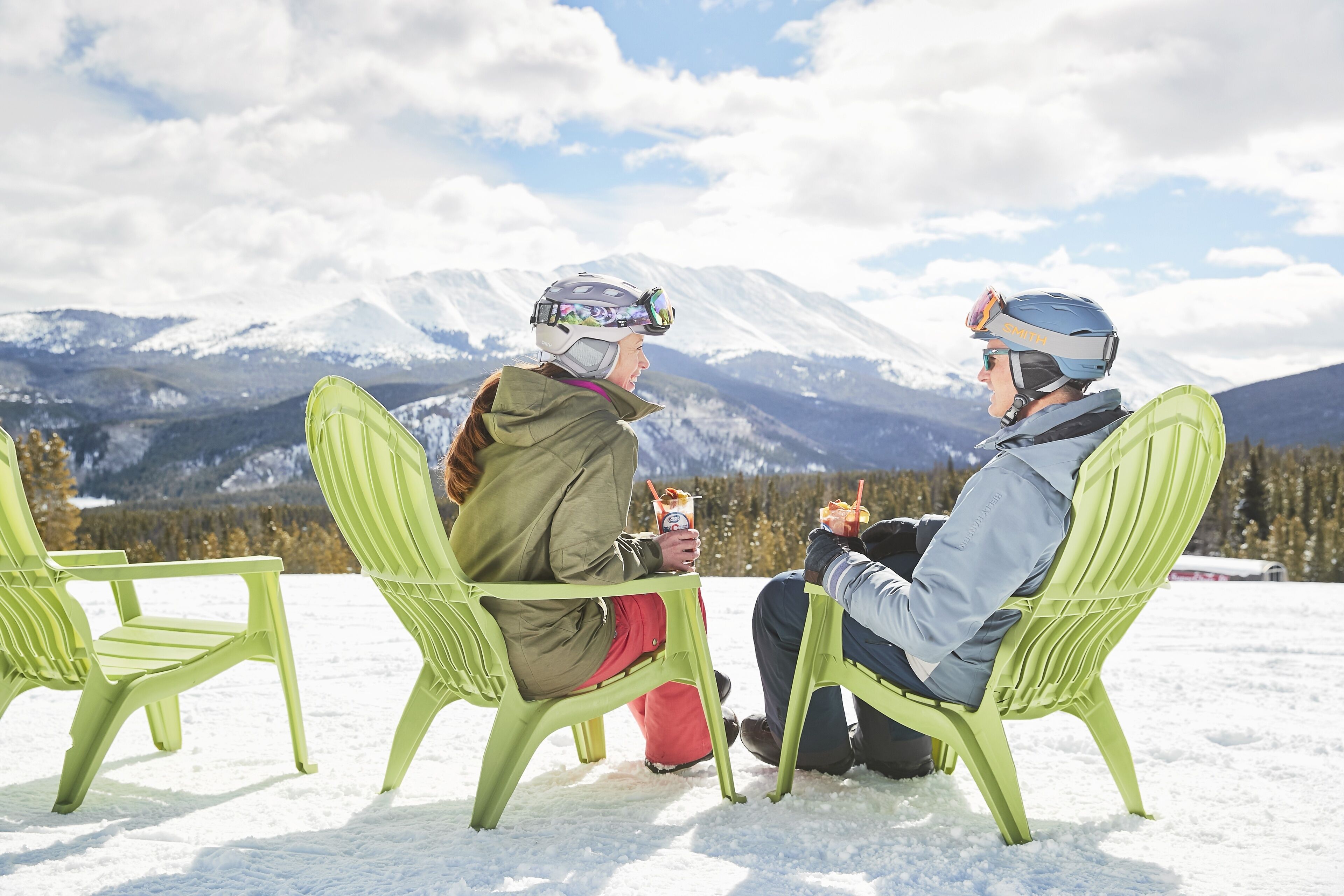 Two people sitting on green chairs outdoors with a snowy mountain backdrop.
