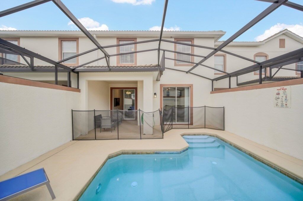 Una piscina con zona cubierta, una silla de descanso y un edificio con ventanas y una puerta al fondo.