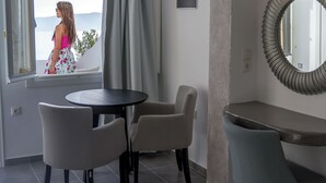 A room with a window overlooking a scenic view, a round table with chairs, and a mirror on the wall. - Remvi Suites (Santorini)