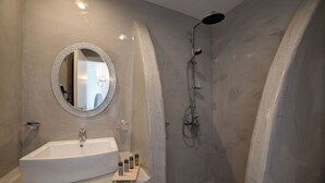 A bathroom with a white sink, a round mirror, and a shower. - Remvi Suites (Santorini)