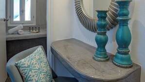 A bathroom with a marble vanity, a large mirror, and a blue patterned chair. - Remvi Suites (Santorini)