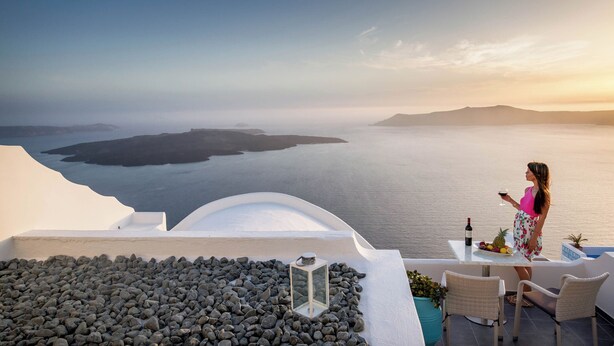 A woman standing on a balcony overlooking a body of water with a sunset in the background. - Remvi Suites (Santorini)