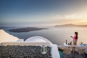 A woman standing on a balcony overlooking a body of water with a sunset in the background. - Remvi Suites (Santorini)