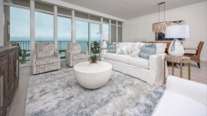 Ein geräumiges Wohnzimmer mit großem Fenster mit Meerblick, ein weißes Sofa mit gemusterten Kissen, ein runder Couchtisch und Korbstühle.