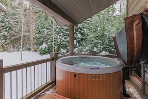 Un bain à remous sur une terrasse en bois, avec une rambarde en métal, entouré d’arbres couverts de neige.