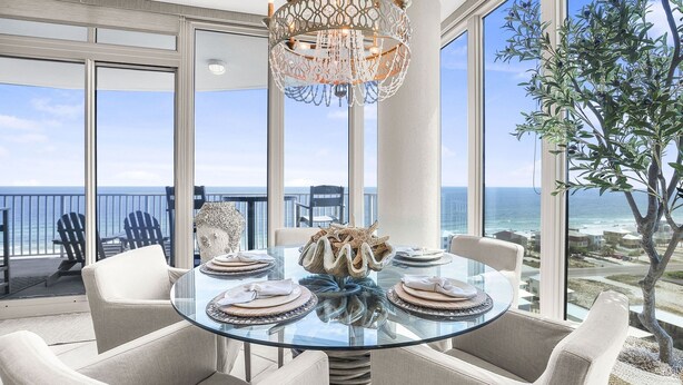 A dining area with a glass table, white upholstered chairs, and a decorative centerpiece. - Lagoon Tower 1301 3 Bedroom Condo (Gulf Shores)