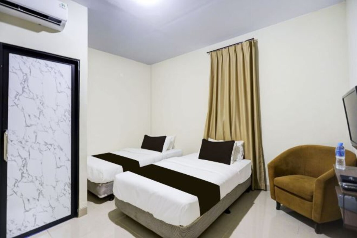 A hotel room with two single beds, a brown armchair, a television, and an air conditioning unit.