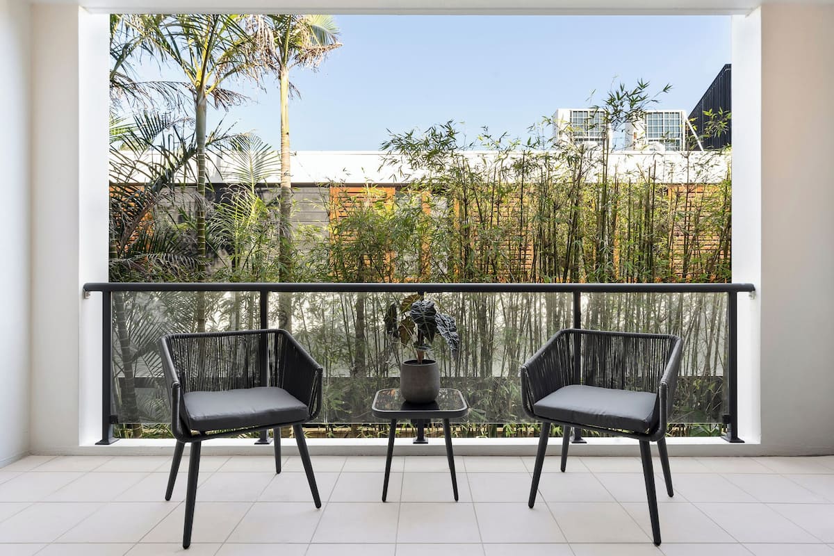 Un balcon avec deux fauteuils en osier noir et une petite table, donnant sur une clôture en bambou et un immeuble.