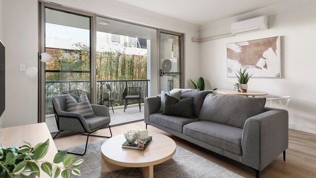 Un salon moderne avec un canapé gris, une table basse ronde et un fauteuil gris. Une porte-fenêtre coulissante donne accès à un balcon extérieur d’où l’on peut admirer une vue sur des bambous et un mur de briques.