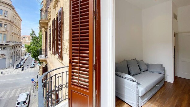 Ein Balkon mit Holzhinterfenstern, einem grauen Sofa und Blick auf eine Straße mit geparkten Autos und Gebäuden.