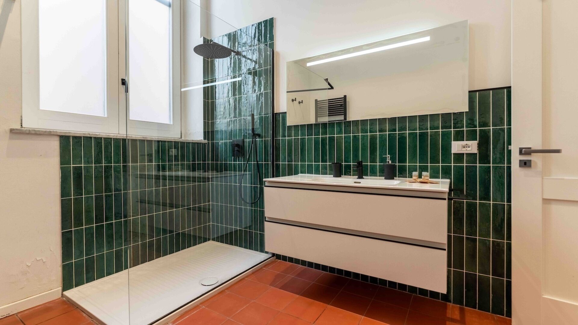 Baño con cabina de ducha de vidrio, tocador blanco con espejo y paredes con azulejos verdes.