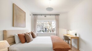 Une chambre à coucher comprenant un lit, une table de chevet, une lampe, un bureau, une chaise et une fenêtre donnant sur des immeubles.