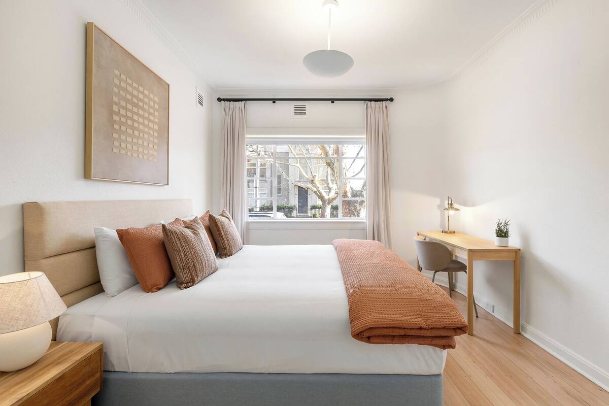 Una camera da letto con un letto, un comodino, una lampada, una scrivania, una sedia e una finestra vista edifici.