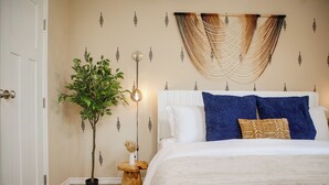 A bedroom with a bed, a potted plant, a bedside table, and a wall-mounted macrame piece.