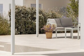 Un área de estar al aire libre con un sofá blanco y una mesa, rodeada de exuberante follaje y una cesta decorativa de flores.