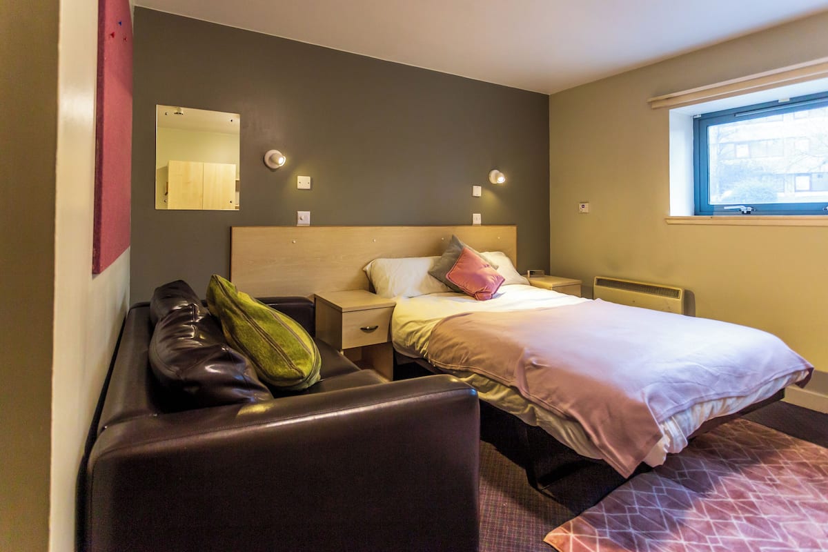 A hotel room with a bed, a nightstand, a chair, a mirror, and a window.