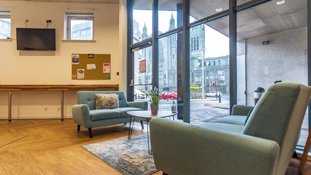 A modern waiting area with a sofa, armchair, and coffee table. A bulletin board and a flat-screen TV are mounted on the wall. - Student Only Pittodrie Street (Aberdeen)