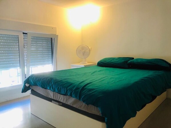 A bedroom with a bed, a nightstand, a fan, and a window with blinds.
