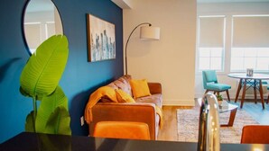 Een moderne woonkamer met een blauwe accentmuur, een grote groene plant, een bank met oranje kussens, een vloerlamp, en een eethoek met een tafel en stoelen.