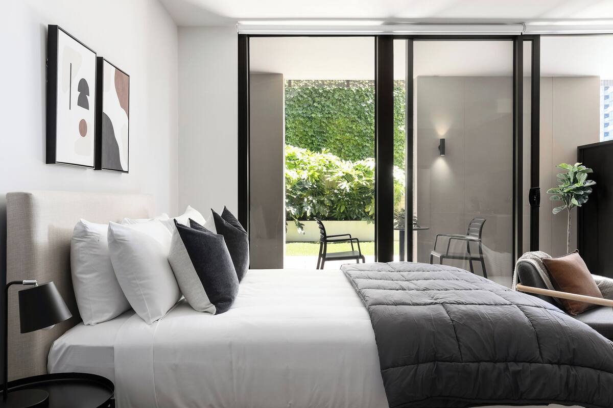 A modern hotel room with a large bed, a desk, and a view of a garden through a sliding door.
