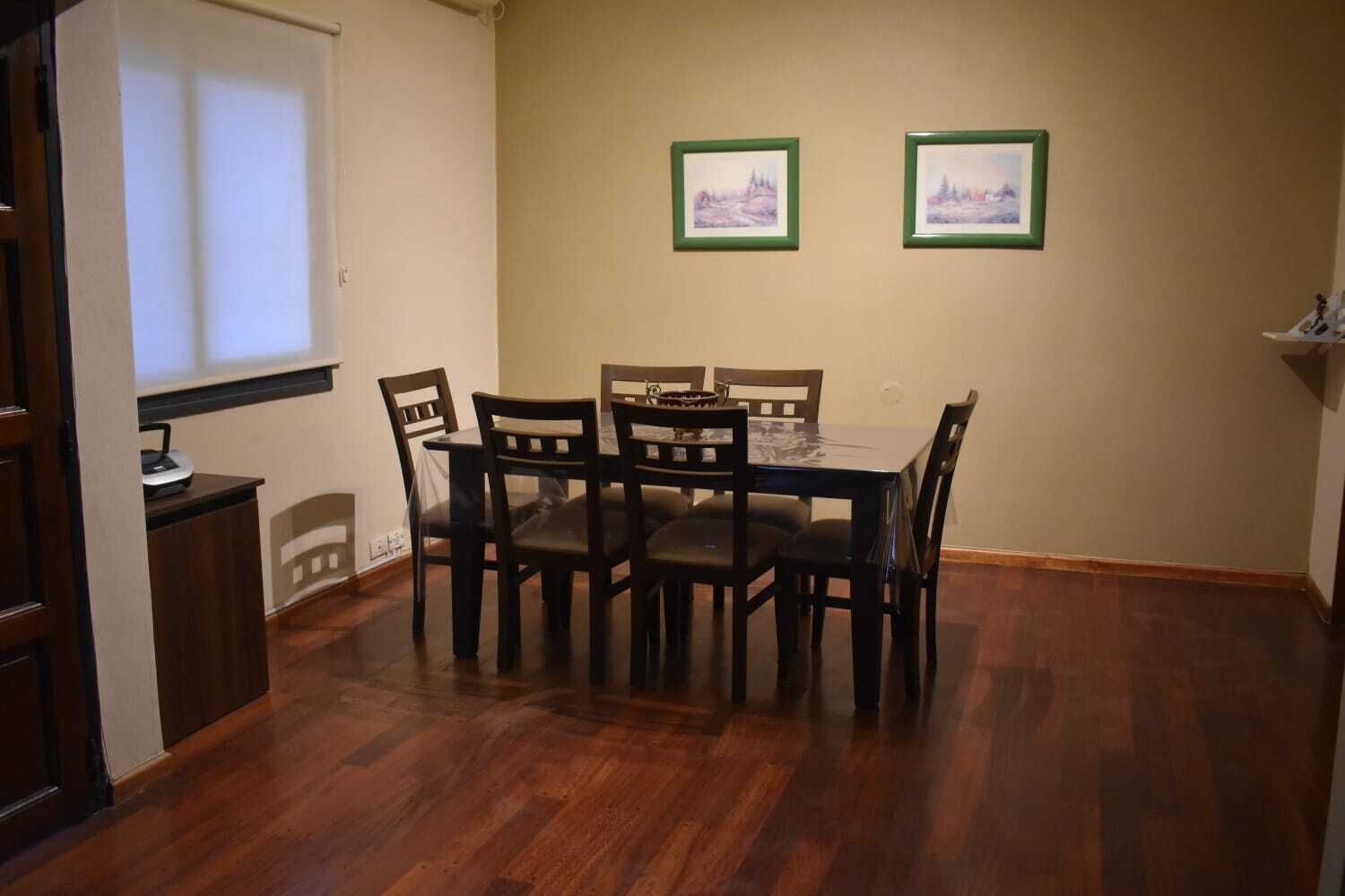 Een eetruimte met een houten tafel en stoelen, twee ingelijste foto’s aan de muur en een raam met jaloezieën.